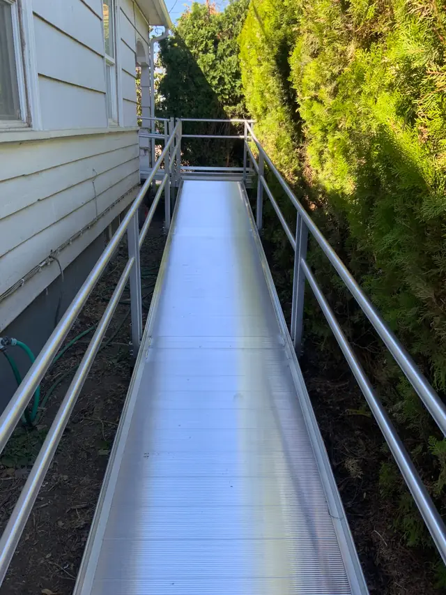 Long Peak Pro aluminum wheelchair ramp with twin handrails running between a house wall and a hedge