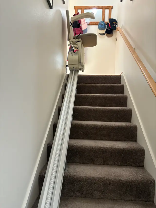 A straight stair lift rail running up a carpeted staircase with the seat parked at the top